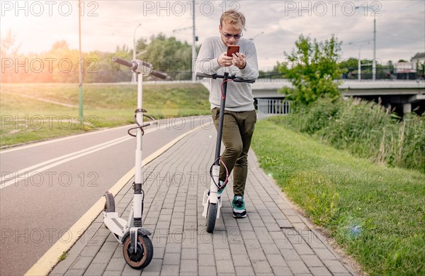 Young handsome blond looking at the route on his smartphone to go with a friend on a trip on electric scooters