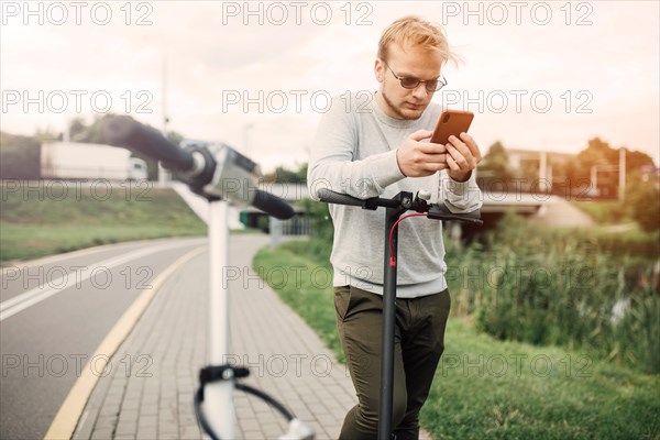 Young handsome blond looking at the route on his smartphone to go with a friend on a trip on electric scooters