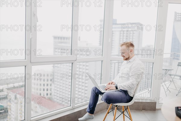 Young IT specialist in a white shirt works at a laptop on a chair in front of a skyscraper window