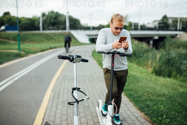 Young handsome blond looking at the route on his smartphone to go with a friend on a trip on electric scooters