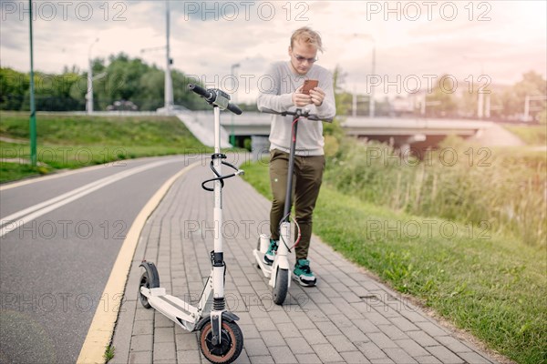 Young handsome blond looking at the route on his smartphone to go with a friend on a trip on electric scooters