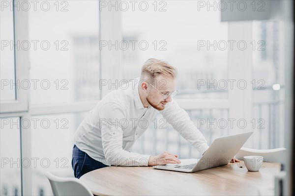 Young IT specialist in a white shirt enthusiastically works at a laptop in front of a skyscraper window