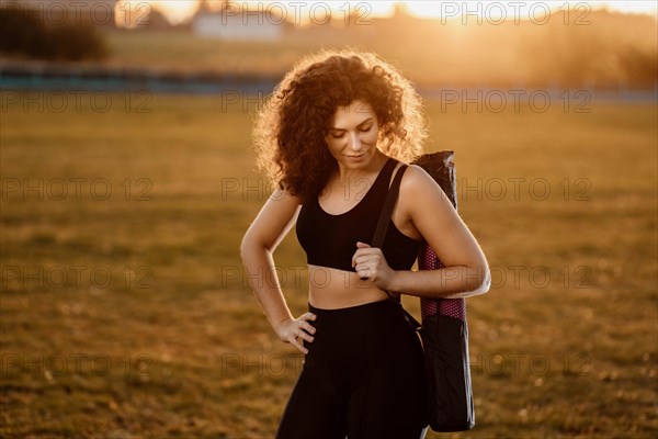 Curly girl runner with a yoga mat stands at sunset on the lawn