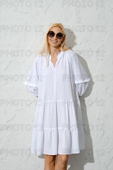 Satisfied blonde woman in sunglasses and white sundress pressed against wall