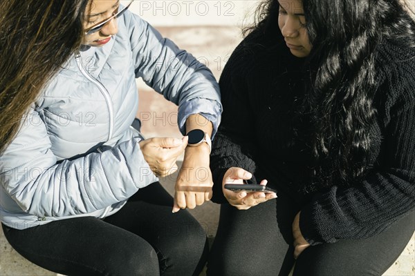 Two beautiful long hair latin women synchronizing data on smart phone and smart watch
