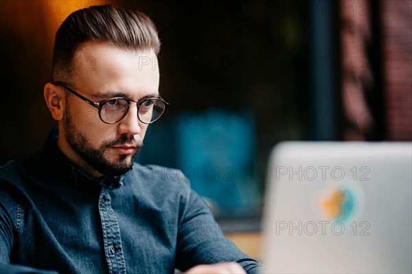 Young successful focused freelancer in a denim shirt working at a laptop in covoking