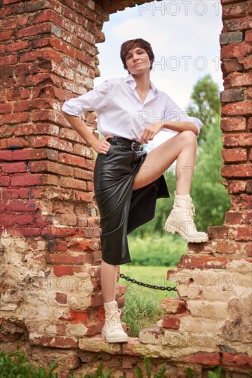 Image of a stylish beautiful woman in a white shirt and leather skirt in a park against the background of a destroyed building. The concept of style and fashion