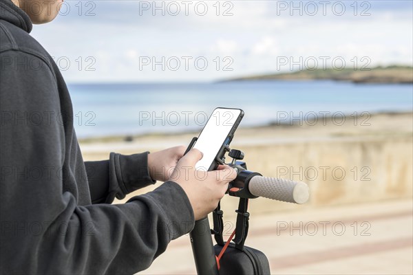 Person holding a smartphone while standing with a scooter by the sea