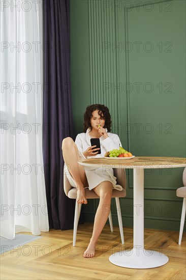 Cute woman in bathrobe sits in living room with smartphone and eats grapes