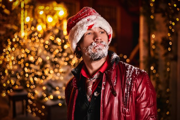 Man dressed as Santa Claus poses on a street decorated with garlands. His beard is covered with snow. New Year and Christmas concept. Mixed media