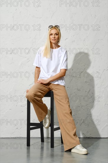 Trendy adult woman in urban-style outfit posing on tall stool in white studio