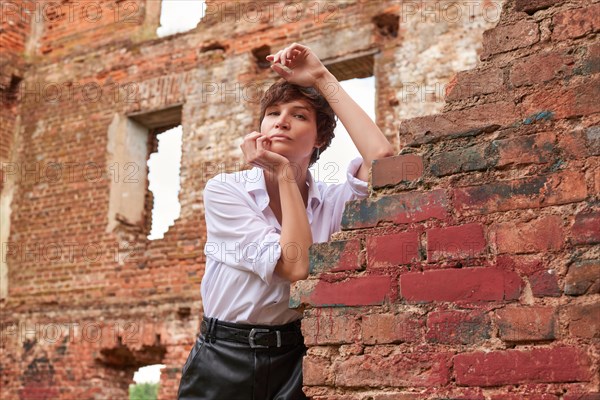 Image of a stylish beautiful woman in a white shirt and leather skirt in a park against the background of a destroyed building. The concept of style and fashion