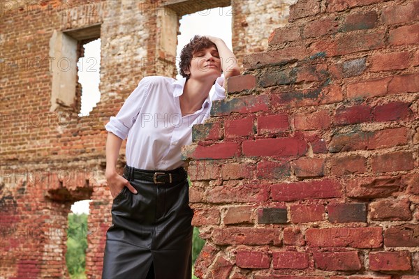 Image of a stylish beautiful woman in a white shirt and leather skirt in a park against the background of a destroyed building. The concept of style and fashion