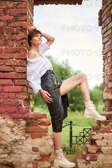 Image of a stylish beautiful woman in a white shirt and leather skirt in a park against the background of a destroyed building. The concept of style and fashion