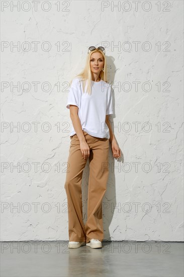 Trendy adult woman in brown wide trousers and white t-shirt leaning on studio wall