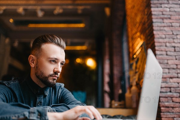 Young successful focused freelancer in a denim shirt working enthusiastically at a laptop in a covoking space