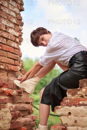 Image of a stylish beautiful woman in a white shirt and leather skirt in a park against the background of a destroyed building. The concept of style and fashion