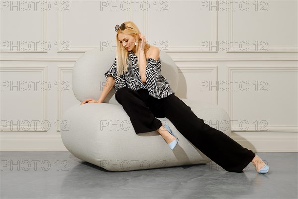 A beautiful blond-haired woman in fashionable boho style clothes sits with her legs crossed in a soft bean bag chair. Her loose trousers and stiletto heels complete the stylish look