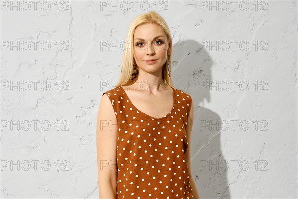 Fashionable blonde woman standing indoors in brown sleeveless blouse with dotted pattern