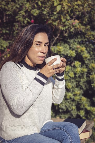 A Latina woman is sitting on a wooden bench in the garden