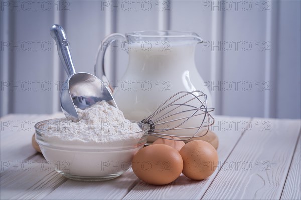 Composition of kitchen objects eggs corolla bowl with flour jug milk on white lacquered old wooden board food and drink concept