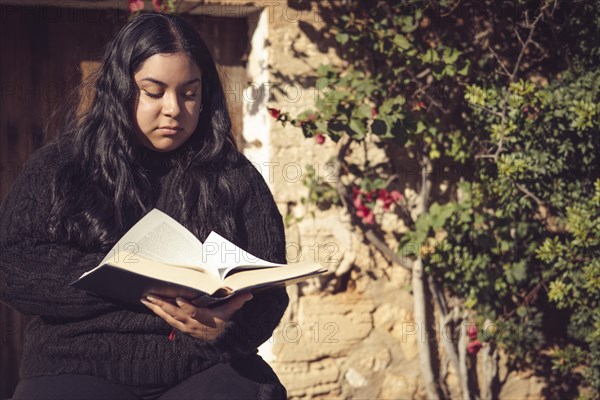 Young latina girl sitting reading a book in the garden next to the country house