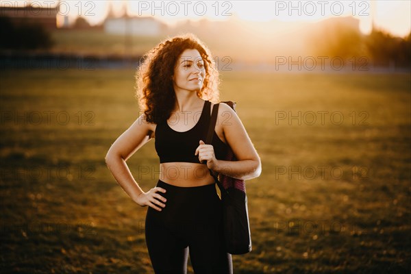 Curly girl runner with a yoga mat stands at sunset on the lawn