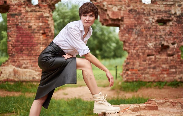 Image of a stylish beautiful woman in a white shirt and leather skirt in a park against the background of a destroyed building. The concept of style and fashion