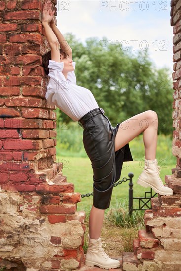 Image of a stylish beautiful woman in a white shirt and leather skirt in a park against the background of a destroyed building. The concept of style and fashion