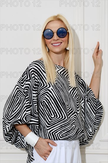 A cheerfully smiling woman in a loose black-and-white patterned blouse and sunglasses leaned her hand against the wall and looked away