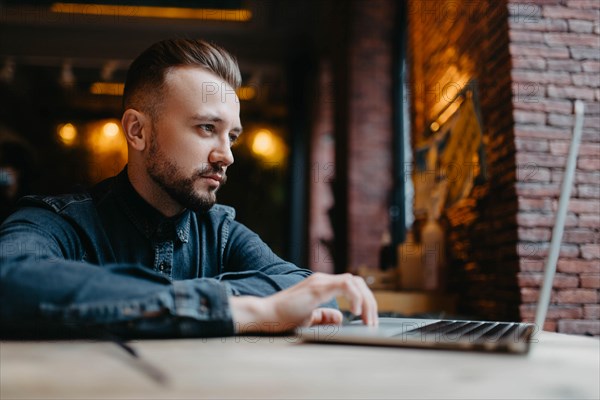 Young successful focused freelancer in a denim shirt working enthusiastically at a laptop in a covoking space