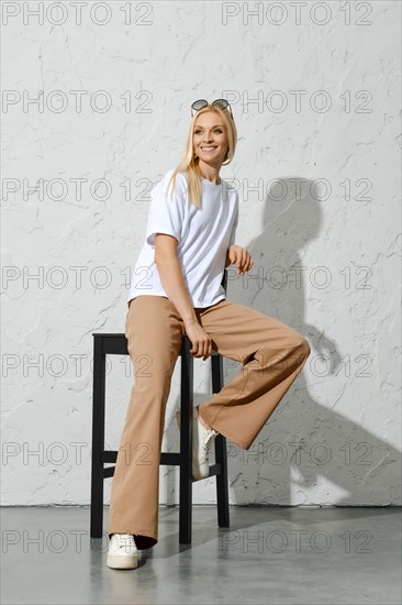 Happy adult woman in urban-style casual clothes sitting on tall stool in white studio