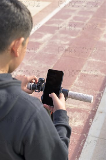 A boy is looking at a smartphone while standing with a scooter on a pathway