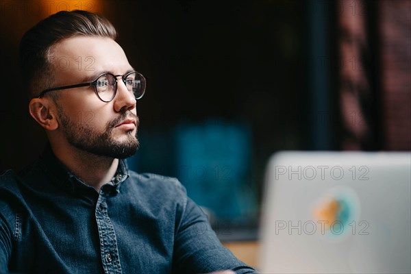 Young successful focused freelancer in a denim shirt working at a laptop in a covoking space and looking away