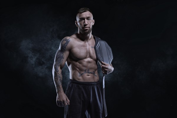 Kickboxer with a belt on his shoulder poses against a background of smoke. Sports competitions. Fight night. The concept of mixed martial arts. MMA