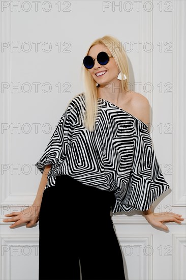 Smiling stylish blonde woman stands indoors wearing a striped blouse and sunglasses