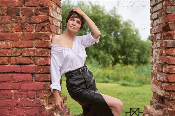 Image of a stylish beautiful woman in a white shirt and leather skirt in a park against the background of a destroyed building. The concept of style and fashion