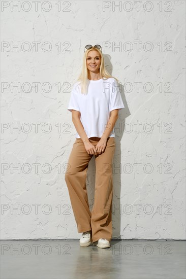 Cute smiling adult woman in oversized trousers and white t-shirt leaning on studio wall