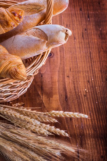 Baguettes and croissants in wicker basket Ears of wheat rye on vintage wooden board Food and drink concept