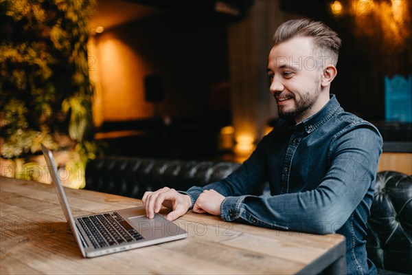 Young successful smiling freelancer in a denim shirt working at a laptop in covoking