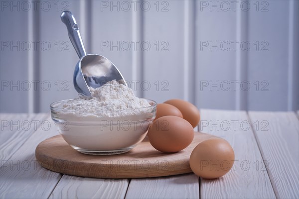 Composition flour in transparent glass bowl scoop eggs on white lacquered old wooden board Food and drink concept