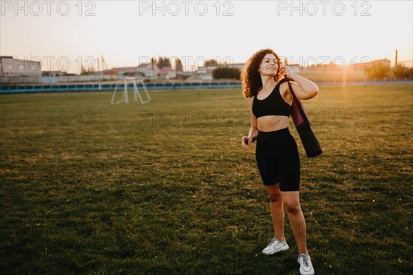 Curly girl runner with a yoga mat stands at sunset on the lawn