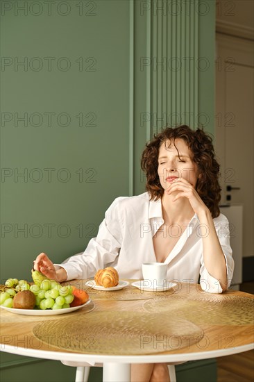 Young woman squints in bright morning sun while sitting at table with coffee and croissant in kitchen