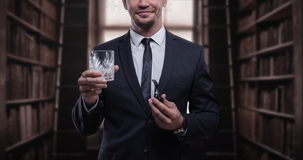 Portrait of an elegant man in a suit with a smoking pipe and a glass in the library. Business concept.