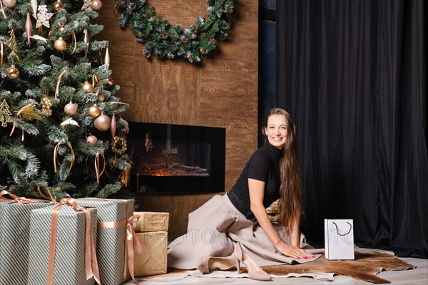 A joyful woman sits on the floor beside a warm fireplace decorated with a lush green wreath. The atmosphere is cozy and intimate