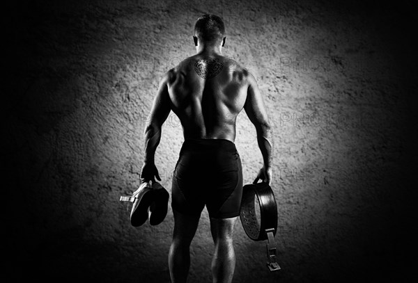 The athlete goes to the gym for training. In his hands he holds sneakers and a belt. Back view