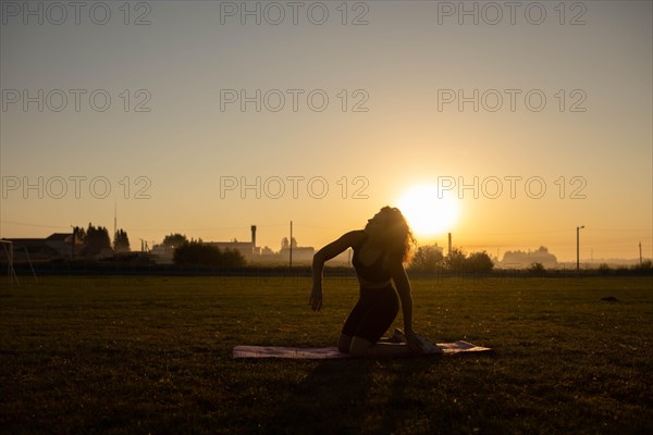 Young curly athletic girl in sportswear doing yoga on a yoga mat outdoors on the grass during sunset