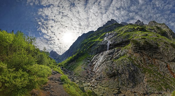 Sunlight breaks through the clouds over a rocky mountain landscape with lush greenery