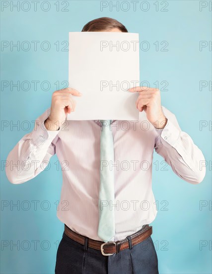 Anonymous businessman covers his head using a blank sheet to hide face expression. Incognito business person hidden behind a white empty paper