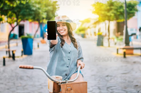 Beautiful girl with hat on bicycle showing cell phone screen on the street. Smiling tourist on bicycle showing cell phone screen on the street